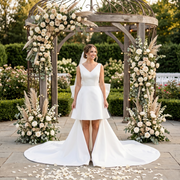 Smiling bride wearing a modern short satin wedding dress with a plunging V-neckline and a dramatic detachable high-low train, standing under a garden gazebo.