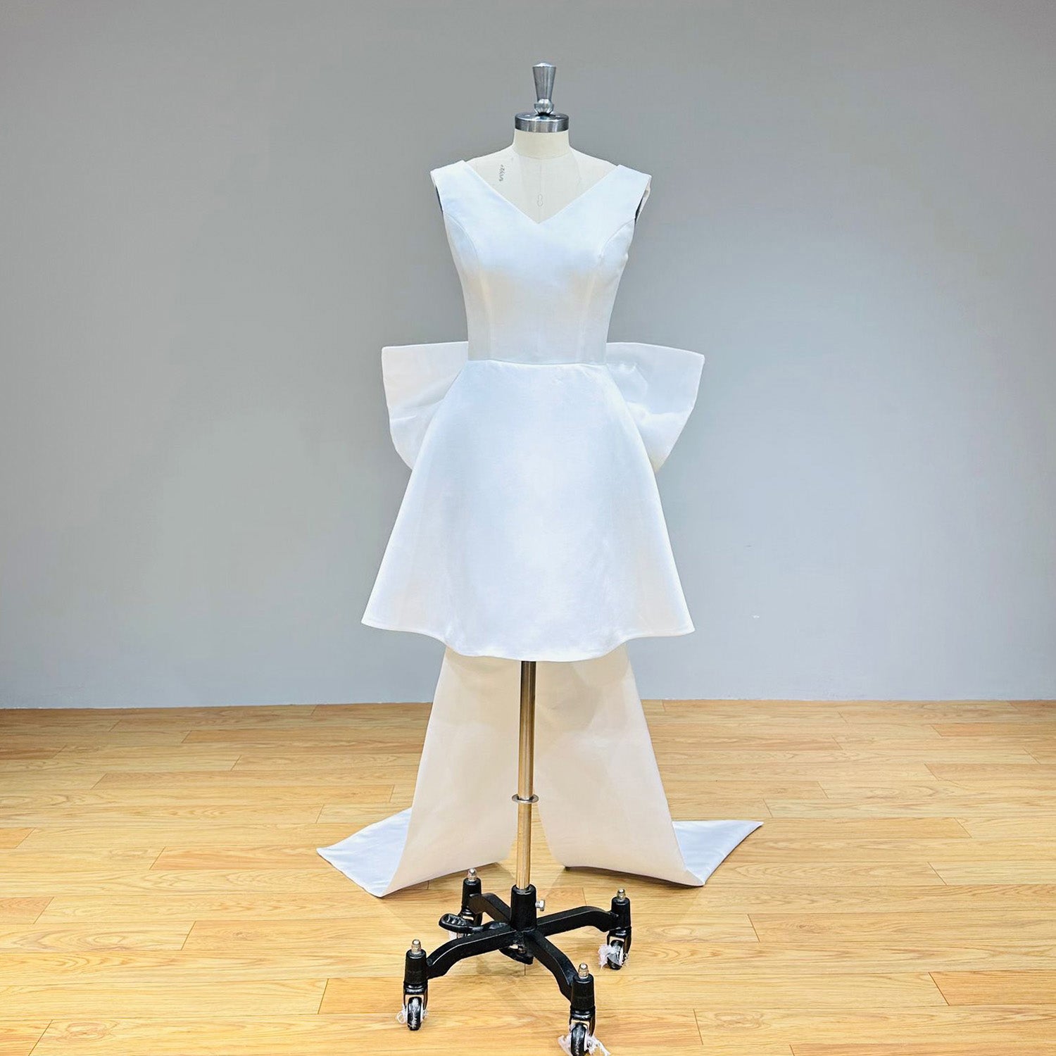 Front view of a minimalist white satin reception dress on a dress form against a solid gray background, highlighting its crisp tailored seams and architectural shape.