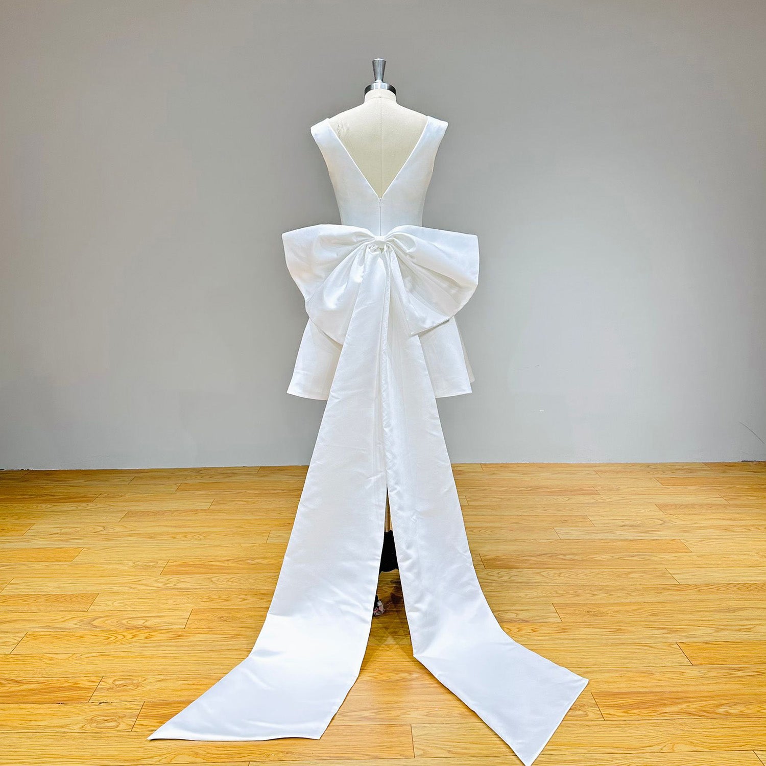 Studio back view of a low V-back short wedding dress on a mannequin, showcasing a spectacular detachable cathedral-length train anchored by a giant satin bow.