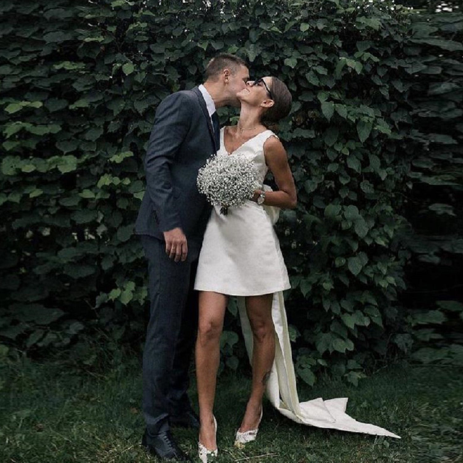 Candid photo of a bride wearing a short white civil wedding dress with a flowing back sash, kissing her groom outdoors while holding a baby's breath bouquet.