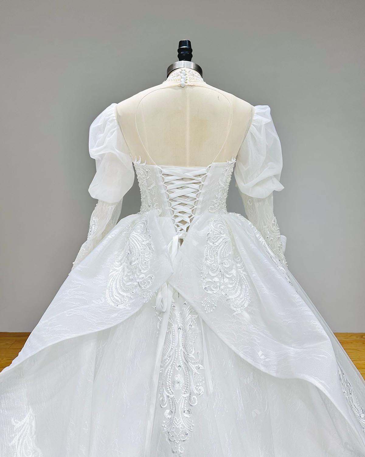 Back view of an elegant high-neck puff sleeve bridal ball gown on a mannequin, featuring a delicate button-back closure and a sweeping lace-trimmed cathedral train.