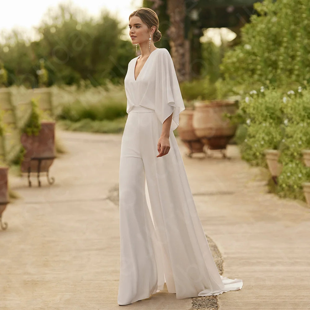 Full-length side profile view of a bride walking outdoors in a holy white wedding pantsuit featuring a deep V-neckline, fluid draping 3/4 kimono sleeves, and flowing wide-leg trousers.