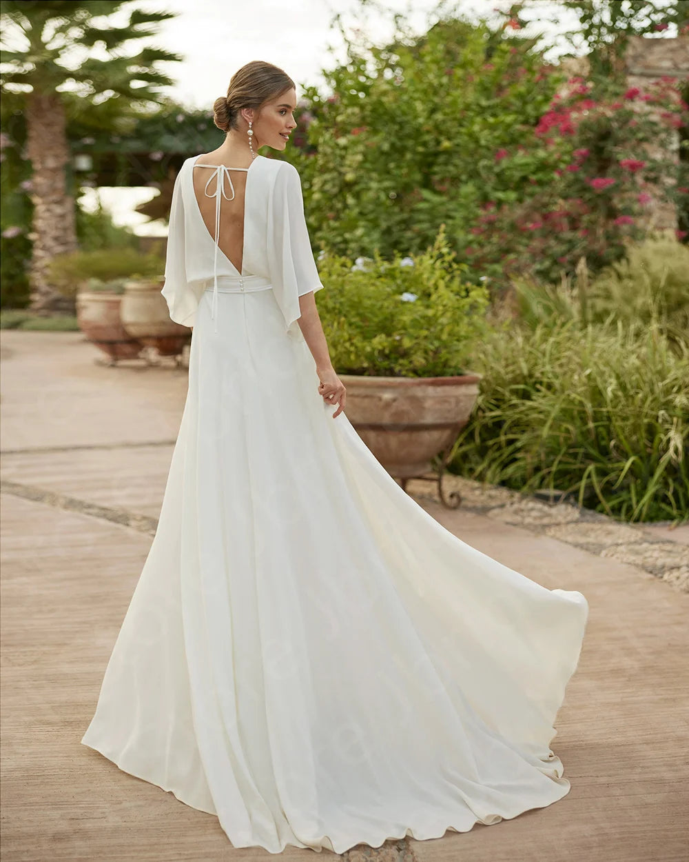 Rear view of an elegant white bridal jumpsuit showcasing a dramatic deep open back with a delicate top tie detail and a sweeping lightweight chiffon train trailing behind.