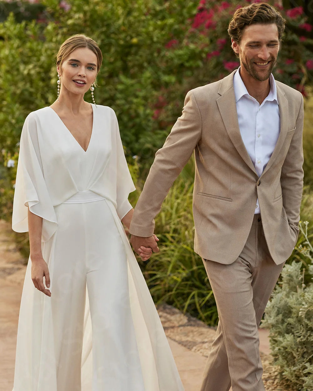 Outdoor wedding photo of a bride wearing a V-neck white wedding pantsuit with loose 3/4 sleeves holding hands with a groom dressed in a tan linen summer suit.