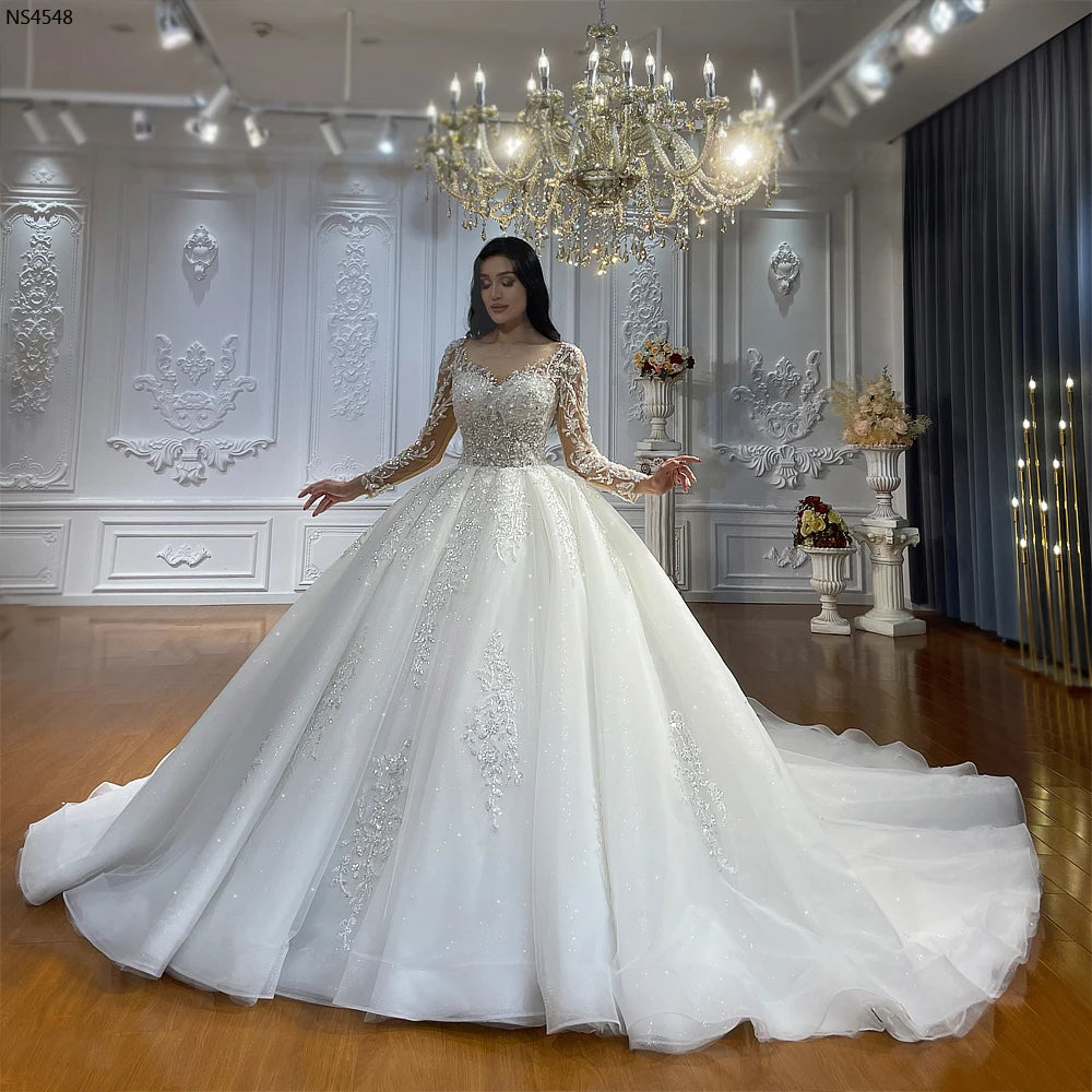 Full-length front view of an elegant model presenting a real sample luxury ball gown bridal dress with a shimmering skirt and long sheer illusion sleeves in a grand showroom.