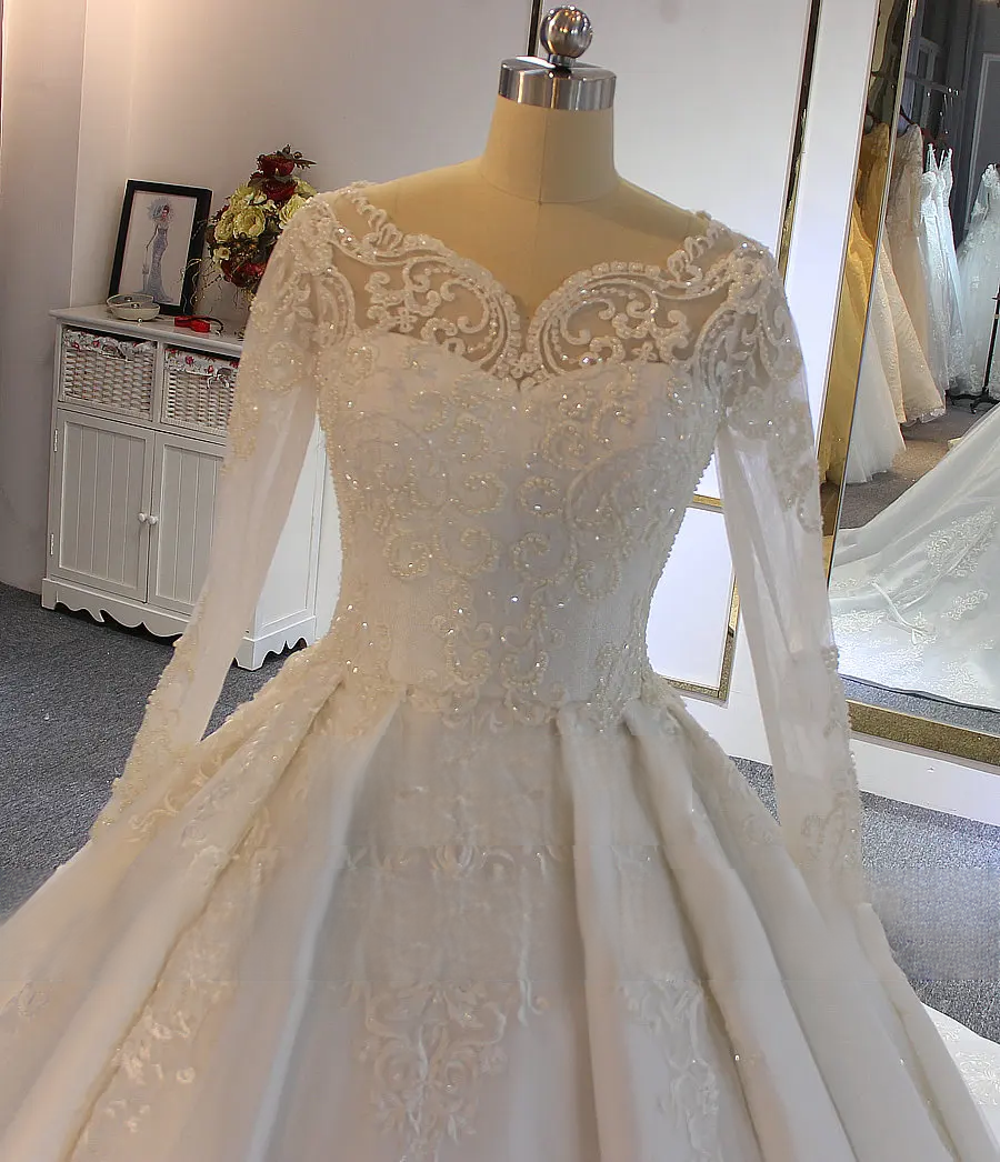 Close-up detail of a bridal gown bodice showcasing a scalloped high neckline, long sheer lace sleeves, and shimmering beadwork.