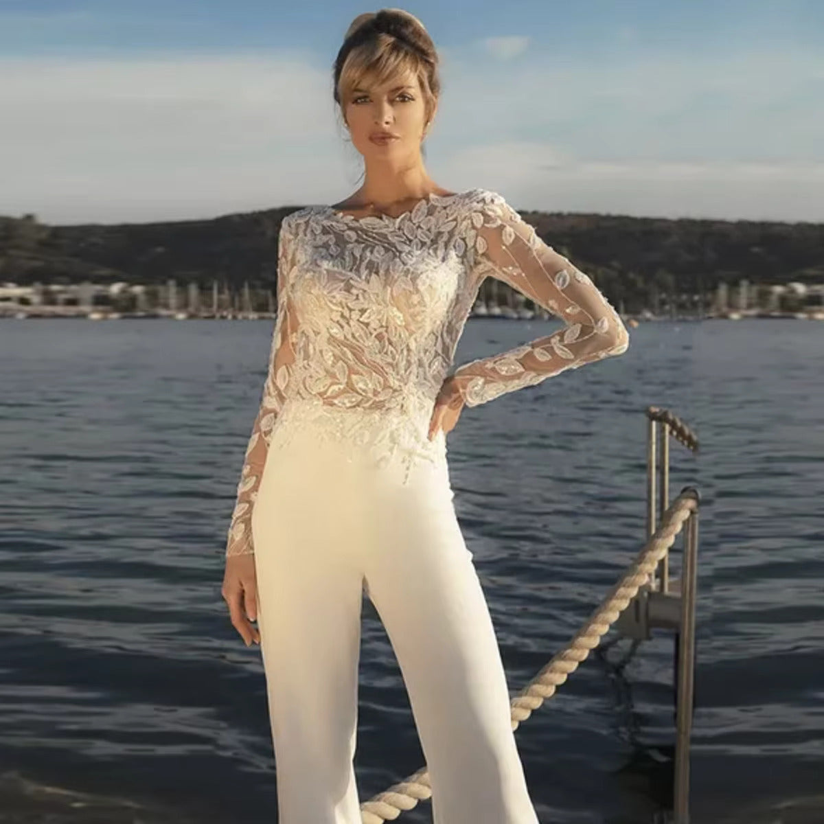 Full-length front view of a bride by the water wearing a modern wedding dress alternative with a sheer illusion lace long-sleeve top and high-waisted white stretch pants.