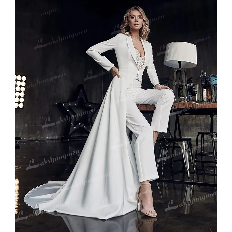 Glamorous portrait of a model sitting on a rustic table wearing a structured long-sleeve white bridal jacket with a dramatic cascading floor-length skirt train over a lace corset pantsuit.