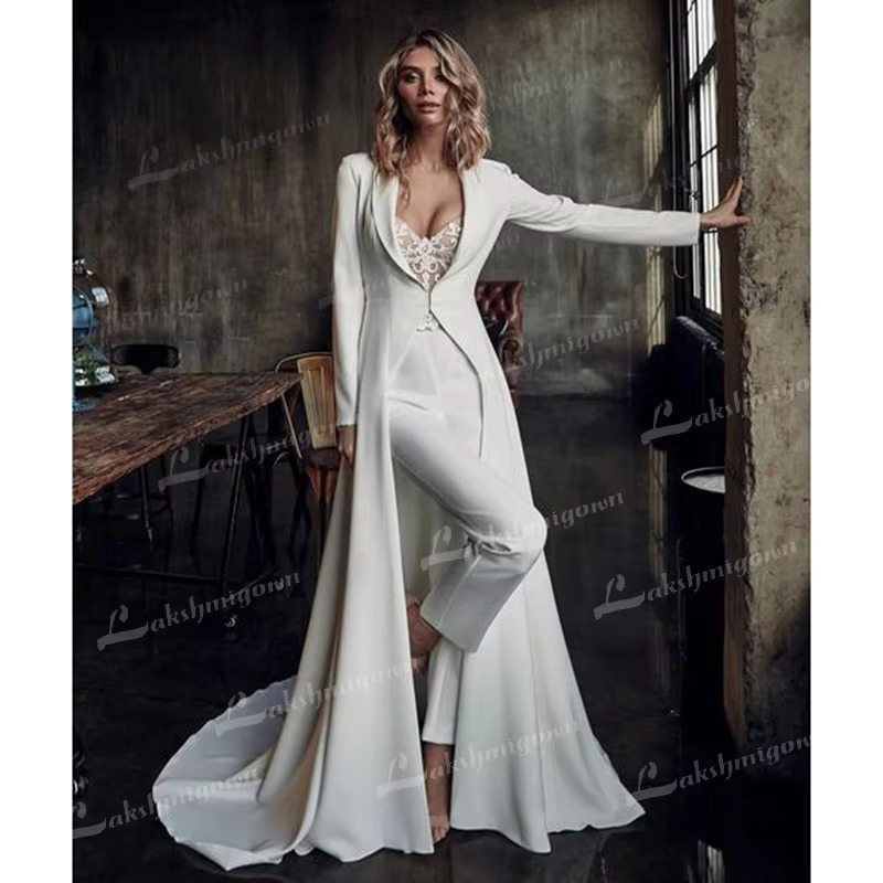 Full-length shot of the alternative bridal gown ensemble, showcasing the fluid sweep of the long-sleeve white wedding jacket train layered over a tailored pantsuit near a window.