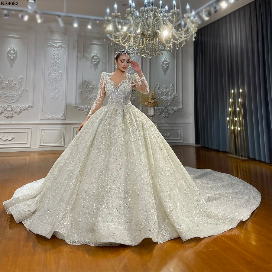 A high-fashion bride wearing a luxury lace appliquéd ball gown wedding dress with long sleeves and a sparkling beaded bodice, featuring an expansive cathedral-length train in an elegant showroom setting.