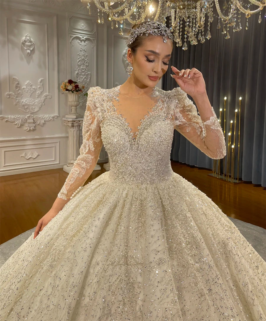 Detailed view of a beaded V-neckline bridal gown featuring intricate lace appliques, long sheer sleeves, and a shimmering crystal headpiece on an elegant model.