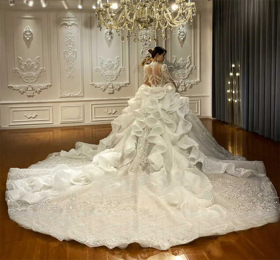 Wide shot of the back of a luxury wedding dress highlighting the cascading tiers of ruffles and a grand lace-embellished royal train with glitter accents.