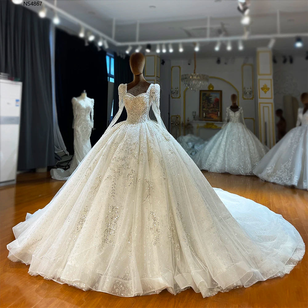 Showroom display of an exquisite full-sleeve ball gown wedding dress on a mannequin, highlighting the regal square neckline and dense, hand-sewn silver beadwork.