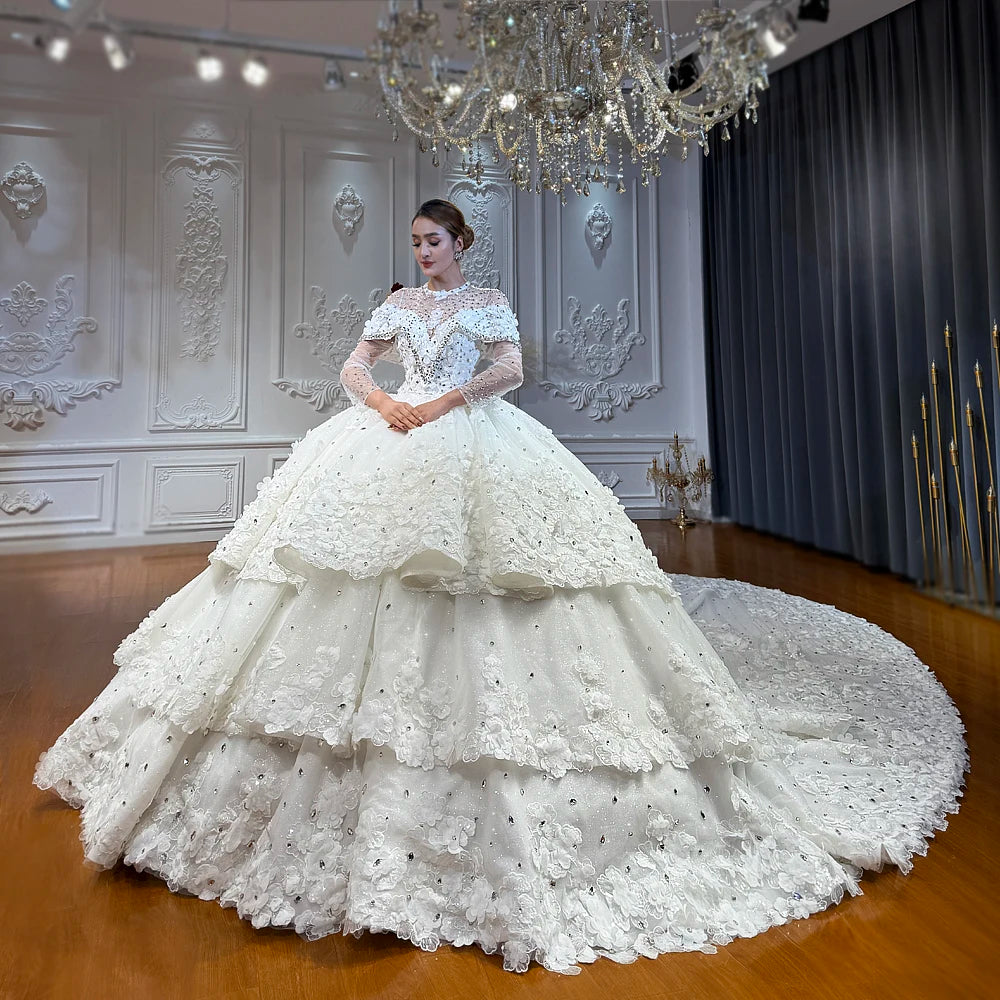 Full-length front view of an elegant model wearing a layered skirt ball gown wedding dress featuring long sheer sleeves, a plunging V-neckline, and intricate 3D lace floral appliqués.
