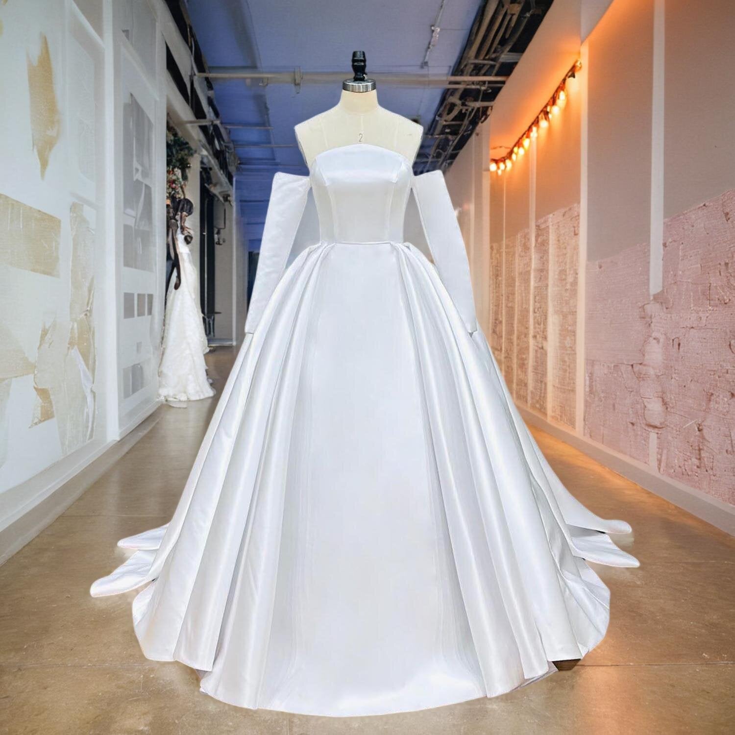 Full front view of a light effect high quality satin ball gown wedding dress on a mannequin in a studio hallway, highlighting the sleek straight neckline, detached long sleeves, and pleated voluminous skirt.