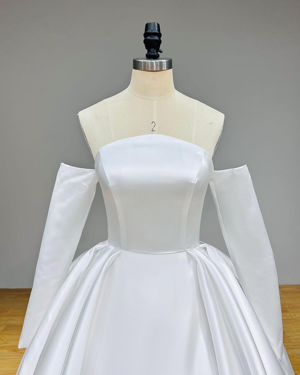 Close-up front bodice detail of a high quality satin wedding dress on a mannequin, emphasizing the minimalist straight across neckline, perfectly fitted waist, and elegant detached long sleeves.
