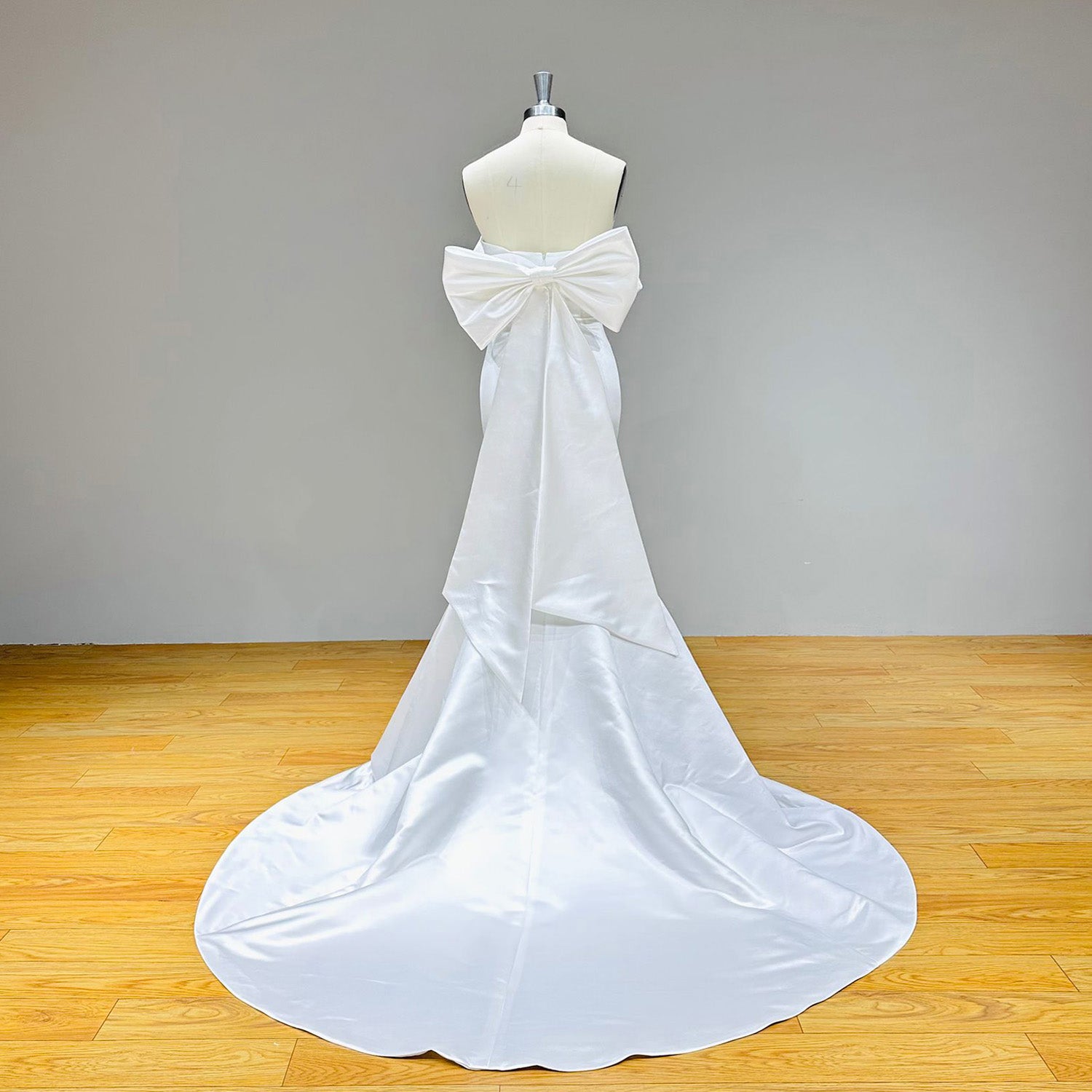 Full-length back view of a smooth satin mermaid wedding gown on a dress form, featuring a spectacular oversized statement bow with long cascading sashes.