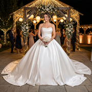 Smiling bride wearing a luxury light effect satin off-shoulder wedding dress with a pleated portrait neckline bodice, standing under a white rose arch during a beach ceremony.