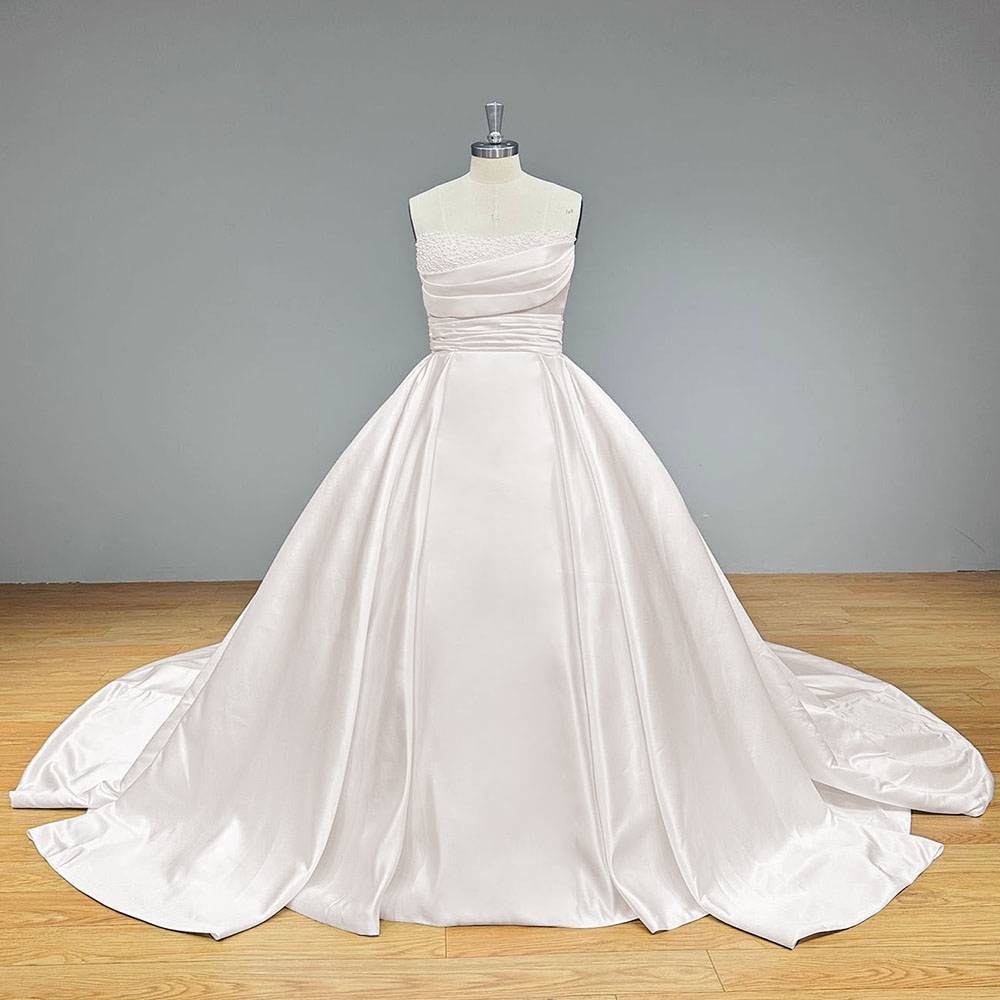 Front view of a minimalist white satin ball gown wedding dress on a dress form, highlighting the clean, light-reflective fabric and symmetrical box pleating at the waist.