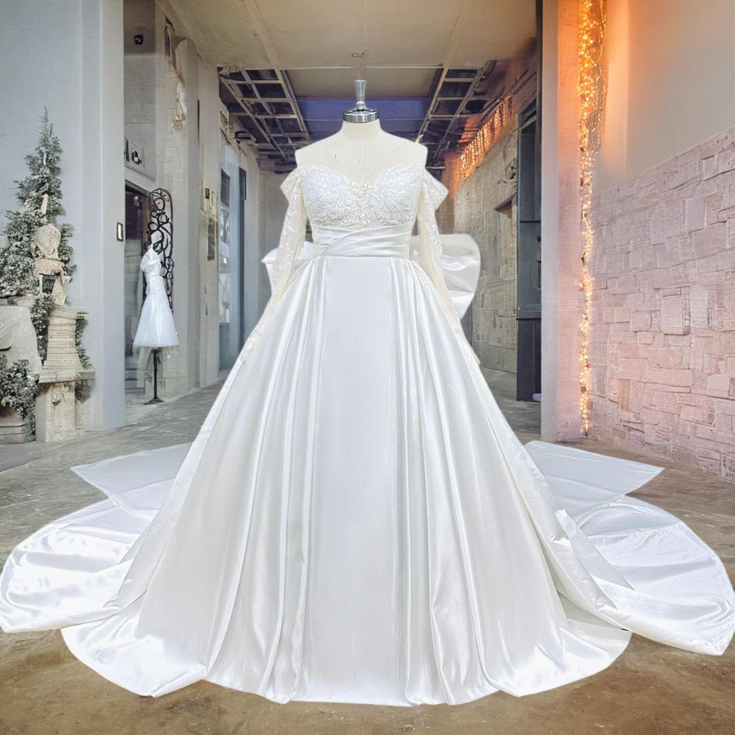 Full front view of a light effect satin wedding dress on a mannequin in a studio hallway, showcasing the off-the-shoulder lace bodice, sheer long sleeves, and expansive chapel train.