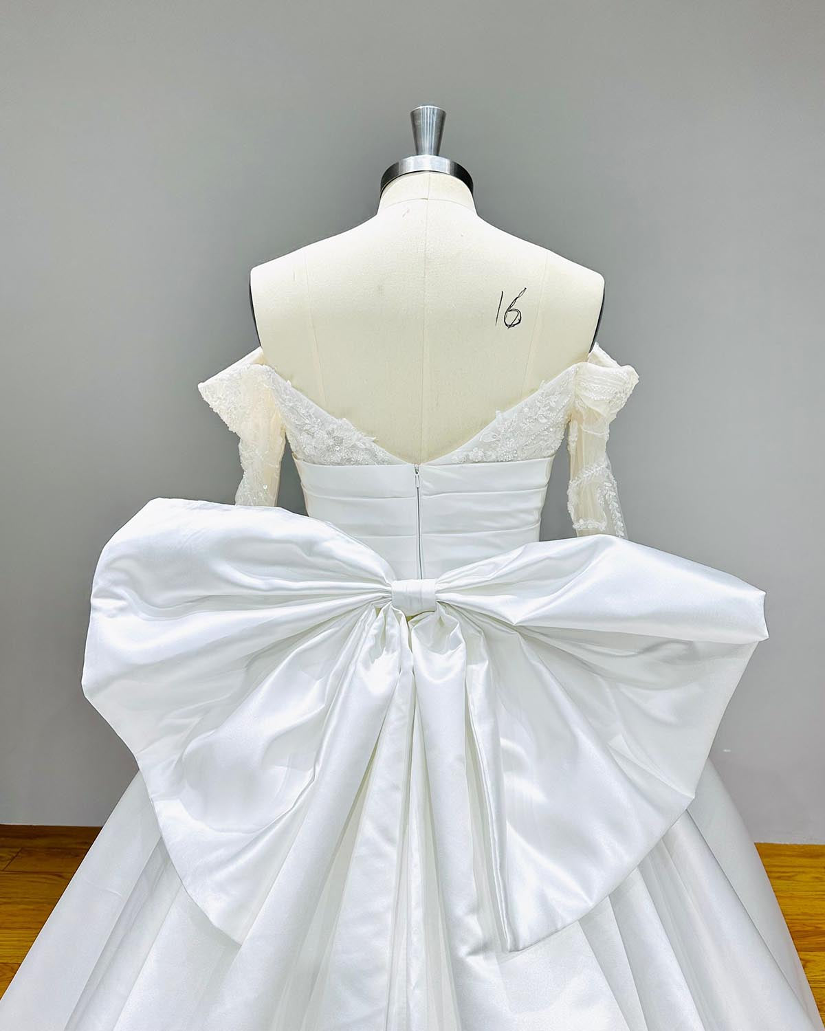 Close-up back view of a light effect satin wedding dress on a mannequin, focusing heavily on the statement detachable big bow at the waistline and delicate off-shoulder lace sleeves.