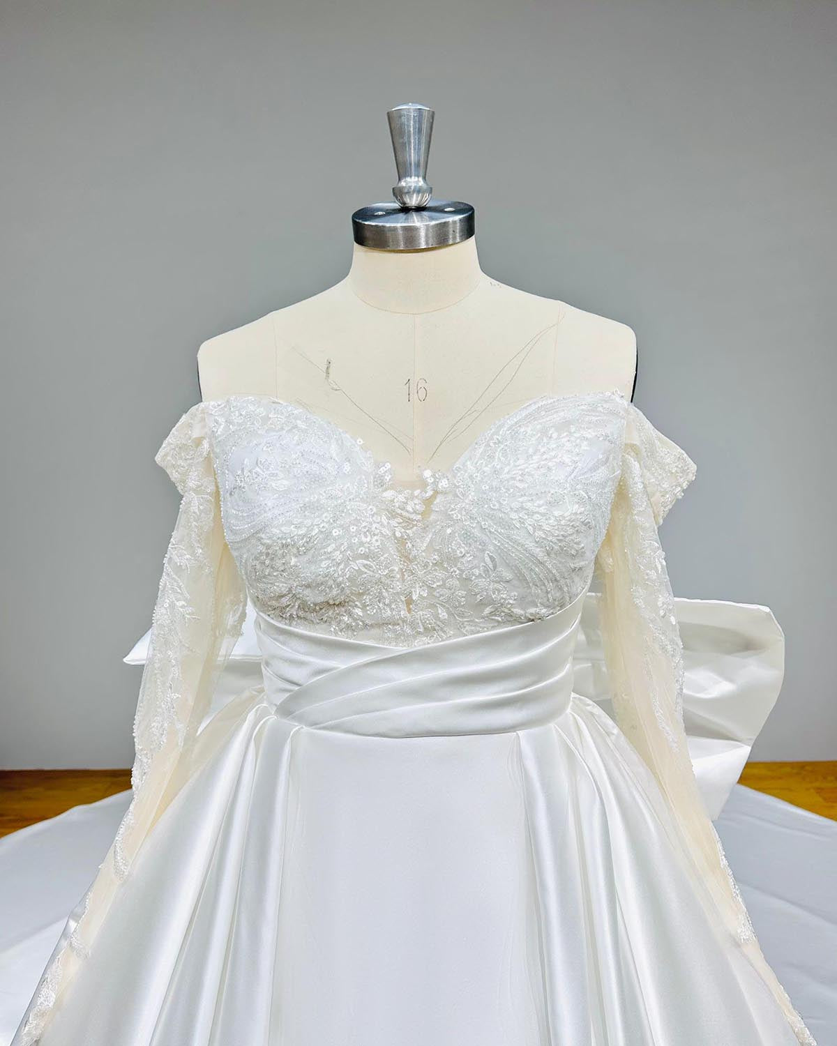 Close-up front bodice detail of a light effect satin wedding dress, emphasizing the intricate sparkling lace sweetheart neckline, off-shoulder sleeves, and pleated satin waistband.