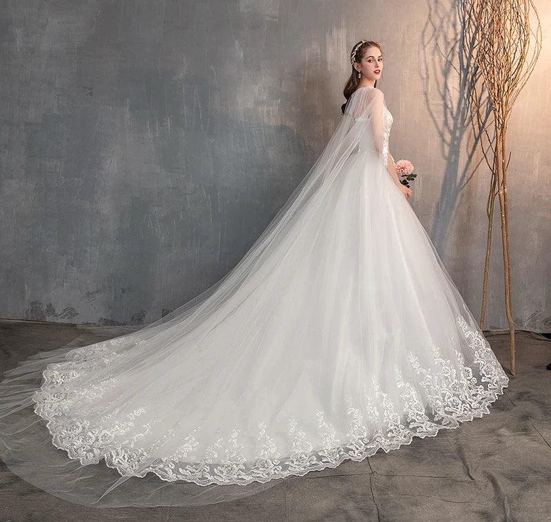 A side profile of a bride in a white wedding gown with a sheer lace cape, showcasing the detailed lace embroidery on the train.