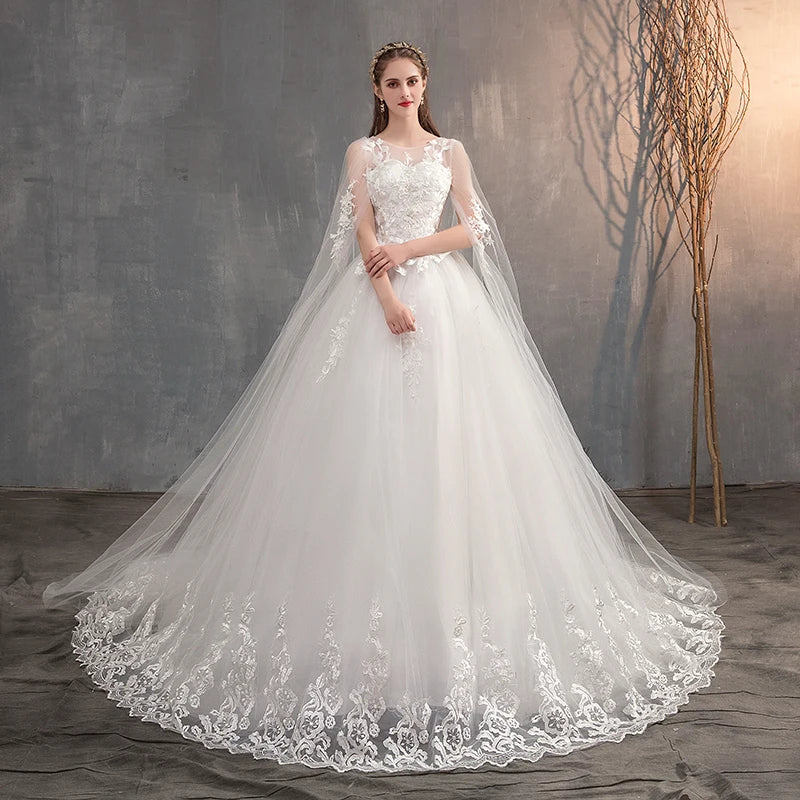 A front-facing portrait of a bride in a white wedding dress with an attached sheer lace cape and ornate embroidery, looking at the camera.