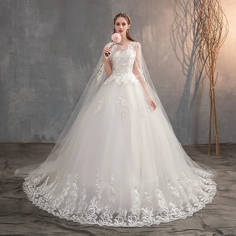 A full-length view of a bride holding a flower, wearing a white wedding gown with a long, sheer lace cape and a detailed train.