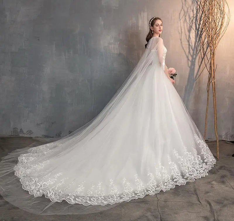 A bride posing in a white wedding dress with a sheer lace cape and a flowing, embroidered train, seen from the side.