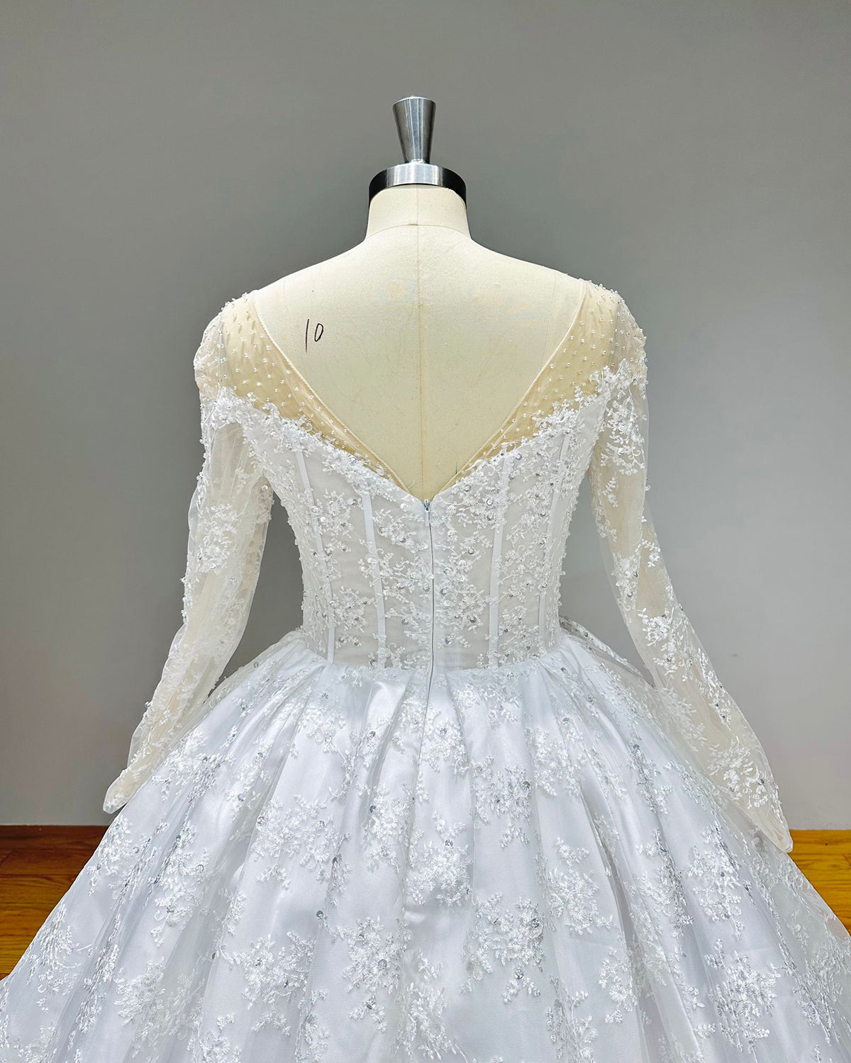 Studio back view of a sheer-top ball gown wedding dress on a dress form, showcasing a delicate button closure along the spine and a wide scalloped lace border on the train.