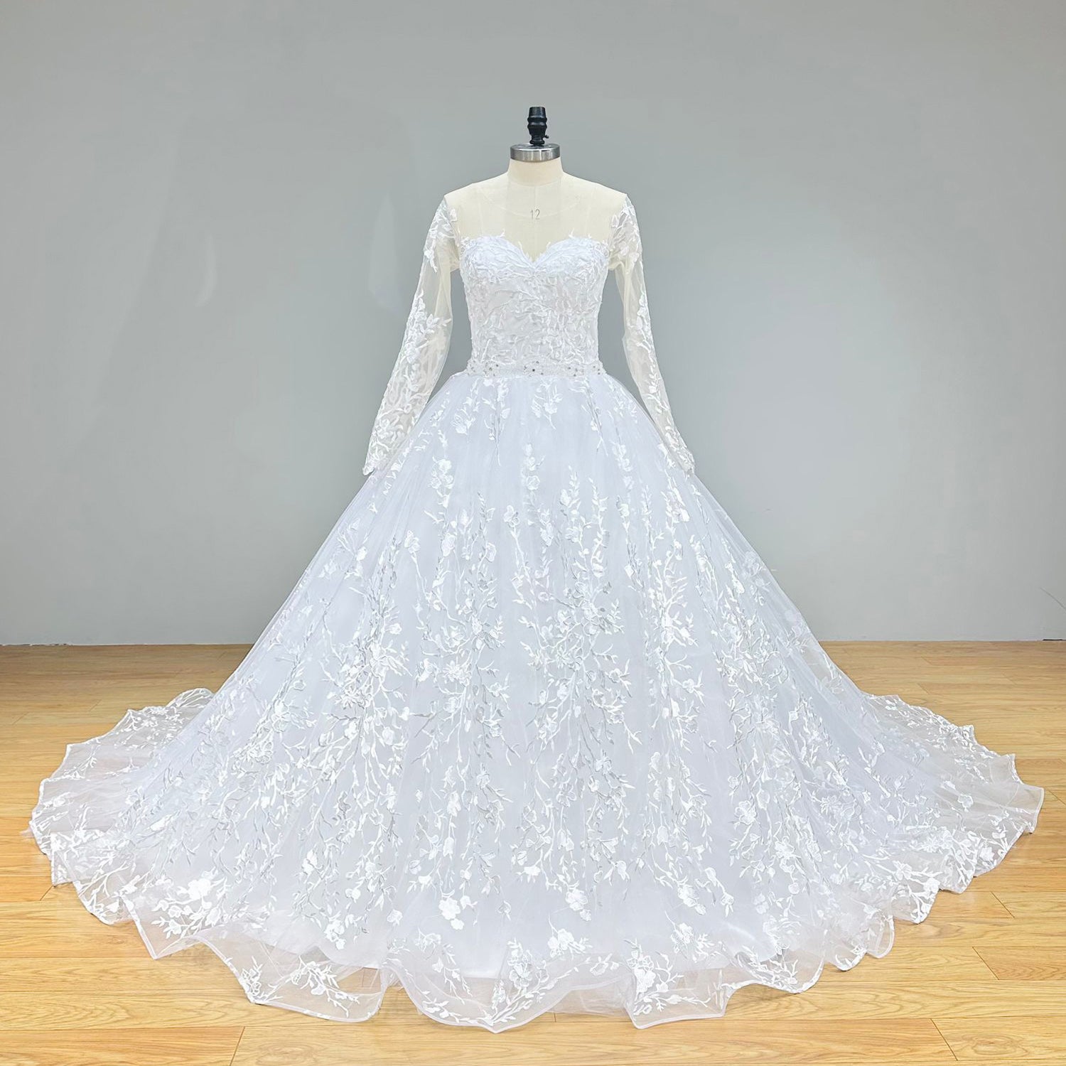 Full front view of a long sleeve ball gown wedding dress on a mannequin in a studio, highlighting the clean, minimalist bodice, cinched crystal beading belt, and dramatic skirt.