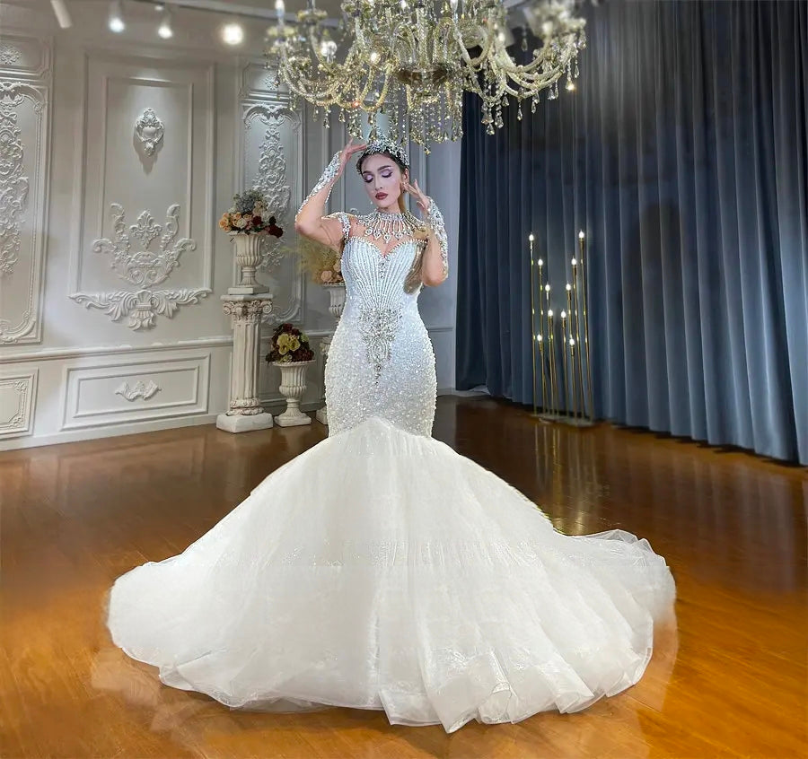 Front-facing view of a stunning beaded mermaid wedding dress featuring sheer long sleeves, intricate pearl embellishments, and a wide-flaring ruffled skirt with a dramatic train.