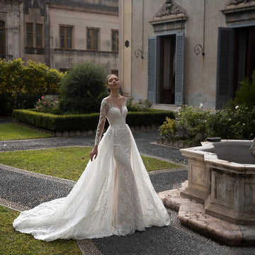 A bride posing in a luxurious long-sleeve mermaid wedding dress, featuring intricate beading, sequins, lace appliques, and a dramatic detachable train.
