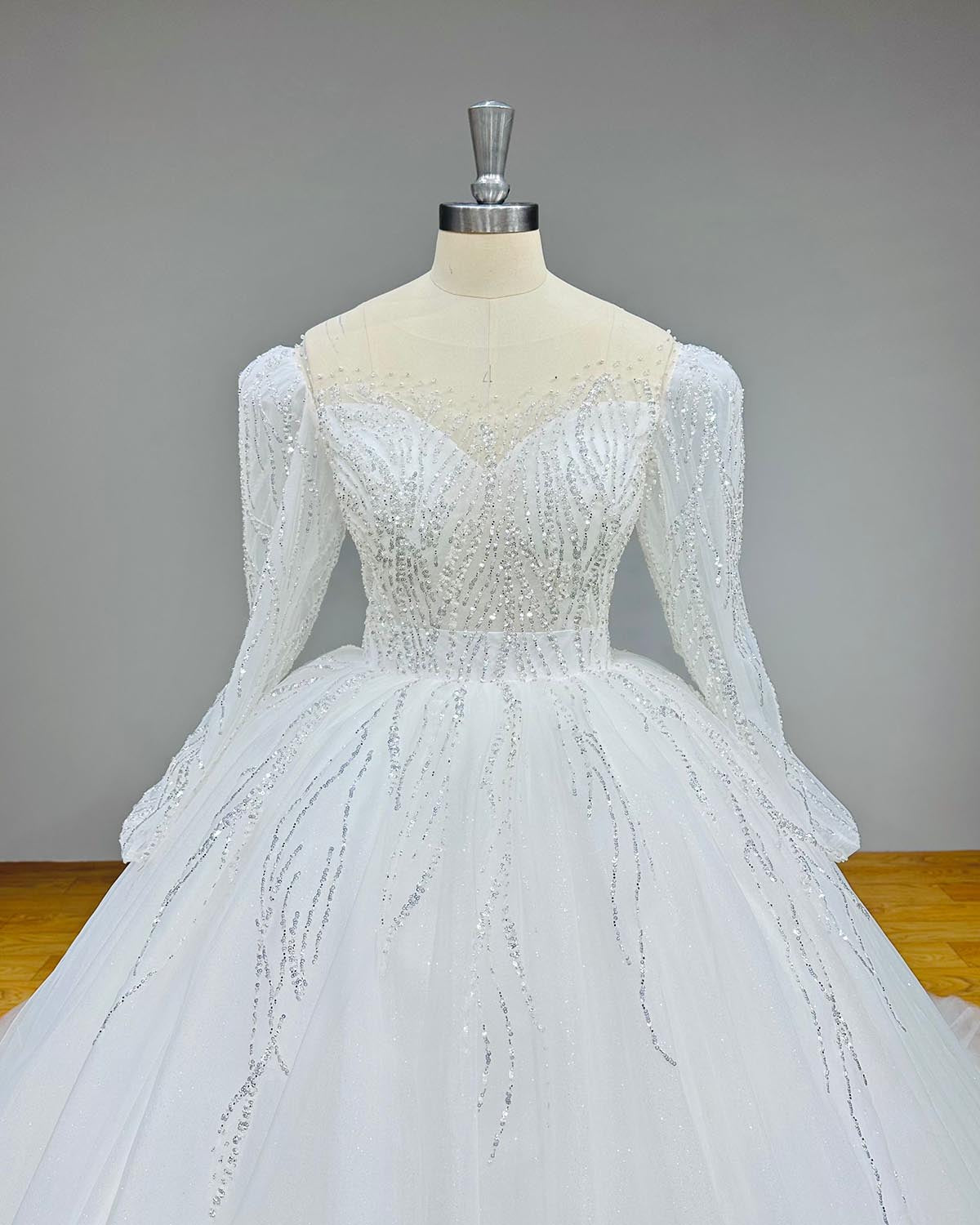 Close-up front bodice view of a luxury long sleeve glitter wedding dress on a mannequin, detailing the sheer illusion sweetheart neckline and delicate linear sequined beading.