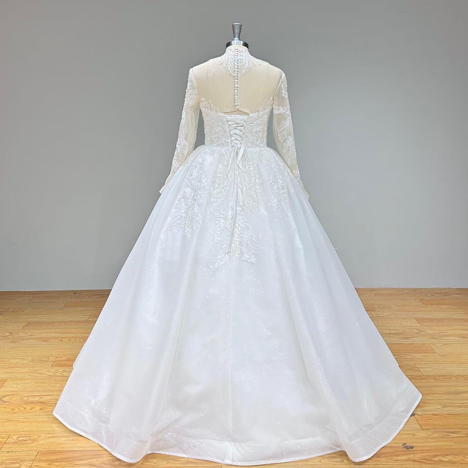Full back view of a long sleeve high neck wedding dress on a mannequin, displaying the sheer illusion back, classic pearl buttons, corset lace-up detail, and shimmering skirt.