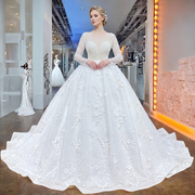 Elegant model posing in a white long sleeve beaded ball gown wedding dress featuring a pearl-encrusted bodice and a voluminous skirt adorned with 3D floral appliqués in a luxury bridal boutique.
