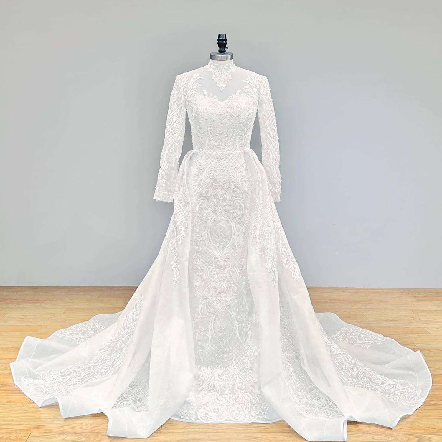 Full front view of a luxury long-sleeve mermaid wedding dress featuring a high illusion neckline, intricate hand-beaded pearl and lace appliqués, and a dramatic detachable train on a display mannequin.