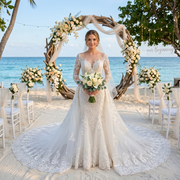 A bride in a vintage luxury long sleeve mermaid wedding dress with a detachable train stands before a driftwood arch at a beach wedding ceremony.