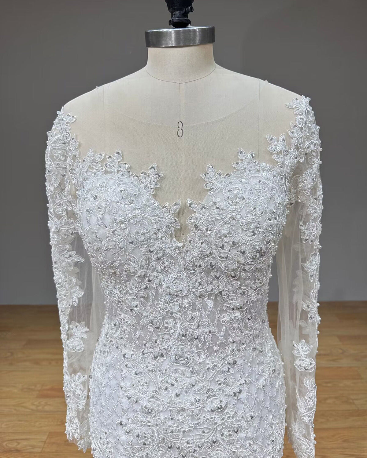 Detailed front close-up of the wedding dress bodice on a dress form, highlighting the sheer illusion plunging V-neckline, scalloped lace edge, and sequin accents.