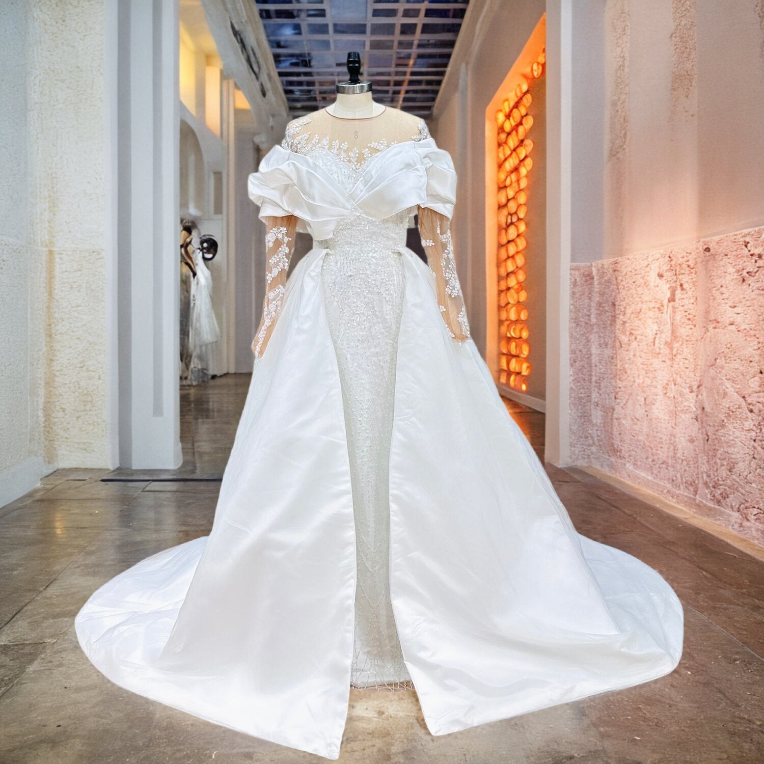 White mermaid bridal gown with an off-the-shoulder portrait wrap neckline, sheer long lace sleeves, and a structured satin overskirt on a mannequin.