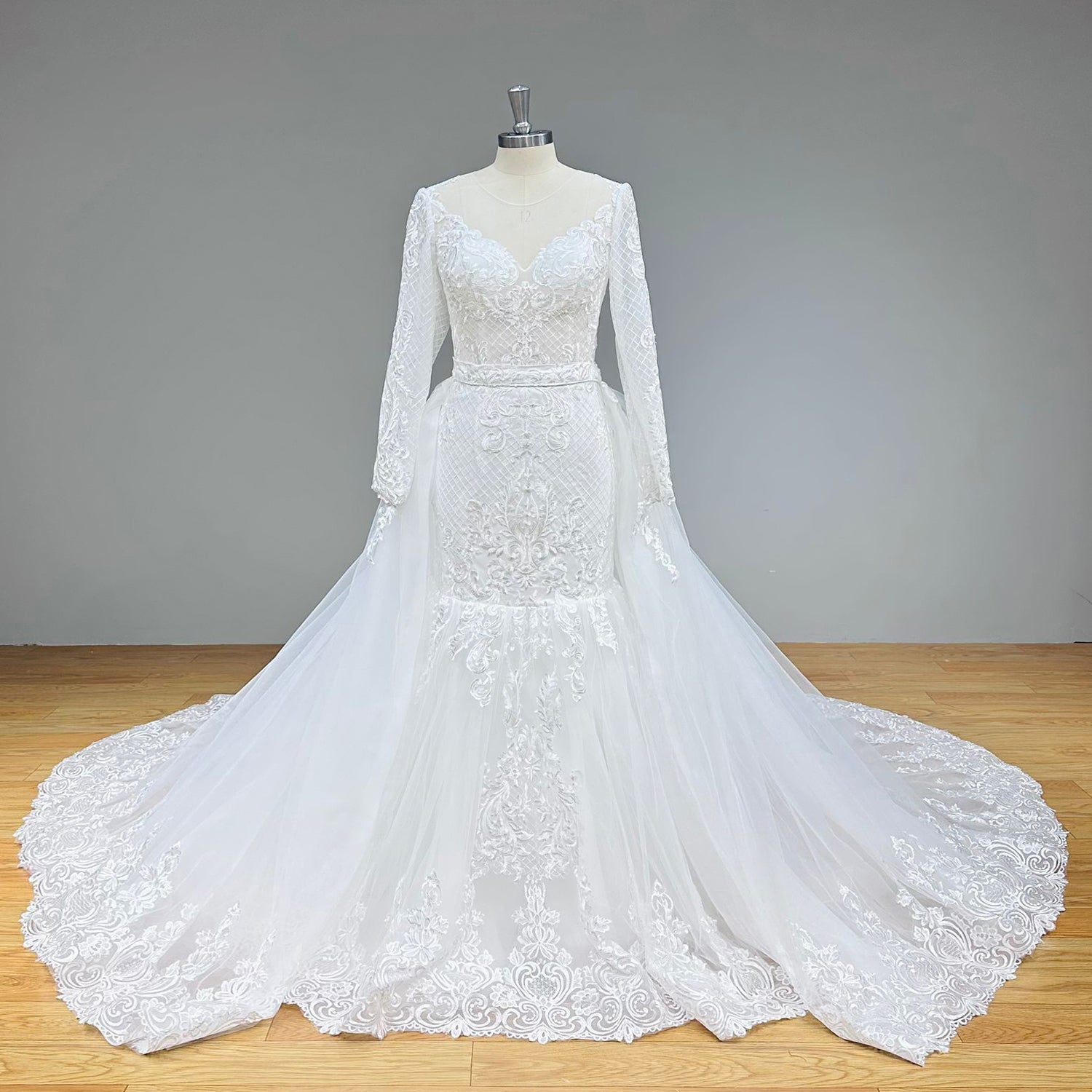 Full front view of a long-sleeve trumpet wedding dress on a mannequin, highlighting the crosshatch lattice embroidery and scalloped lace hemline of the attached overskirt.