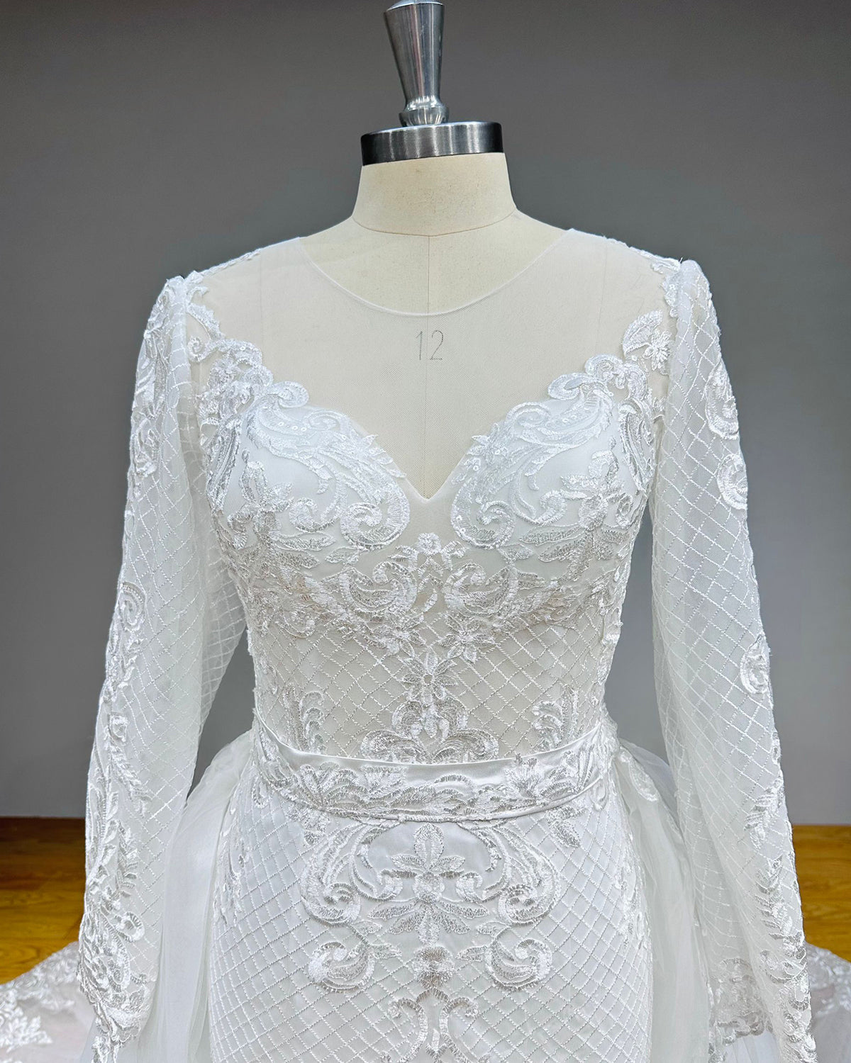 Close-up front view of a bridal gown bodice featuring a sheer illusion neckline, ornate baroque lace appliques, and a textured geometric lattice pattern on the long sleeves.