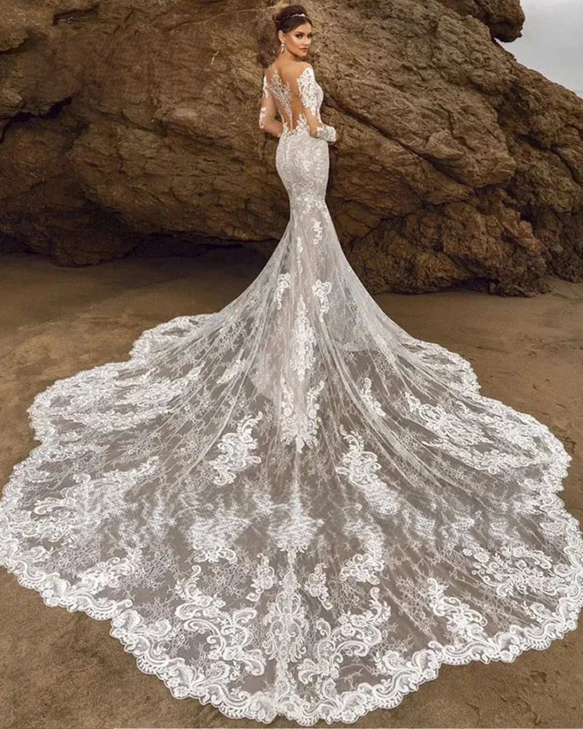 Rear view of a bride on a beach posing in a backless mermaid wedding dress with sheer lace sleeves and a grand cathedral-length lace train.