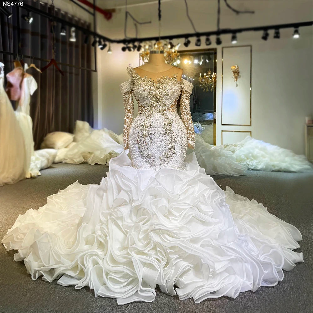 Front view of a luxury long sleeve mermaid wedding dress on a mannequin, featuring heavy 3D crystal embellishments and a voluminous white ruffled skirt in a bridal boutique.