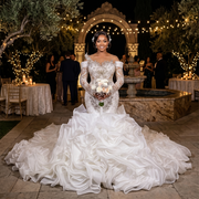 A bride wearing a long-sleeve mermaid wedding dress with elaborate 3D crystal embellishments and a dramatic tiered ruffle skirt at an evening outdoor reception.