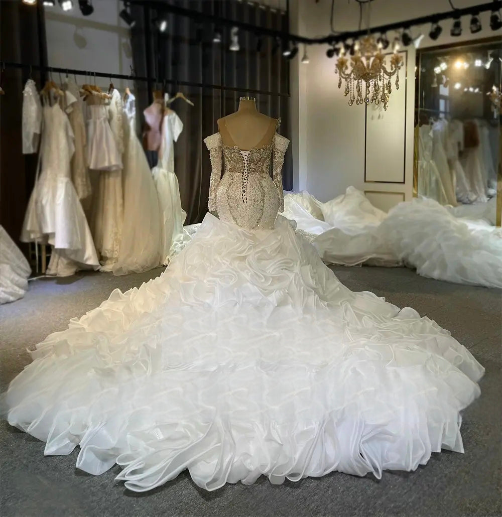 Rear perspective of a long sleeve mermaid bridal dress on a mannequin, detailing the elegant V-back with lace-up corset closure and the cascading white ruffled train.