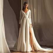 Minimalist long sleeve satin wedding dress with a draped faux-wrap surplice V-neckline, crystal pearl belt, and a dramatic high thigh slit skirt.