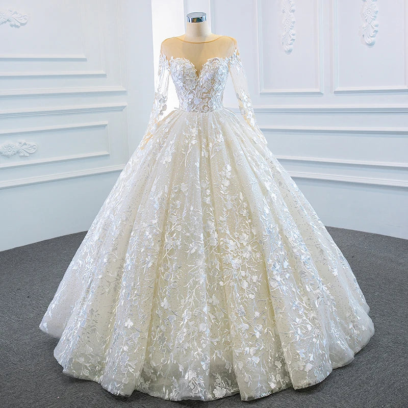 Front full-length view of a luxury long sleeve ball gown wedding dress featuring a transparent illusion neckline and all-over white floral lace appliques.