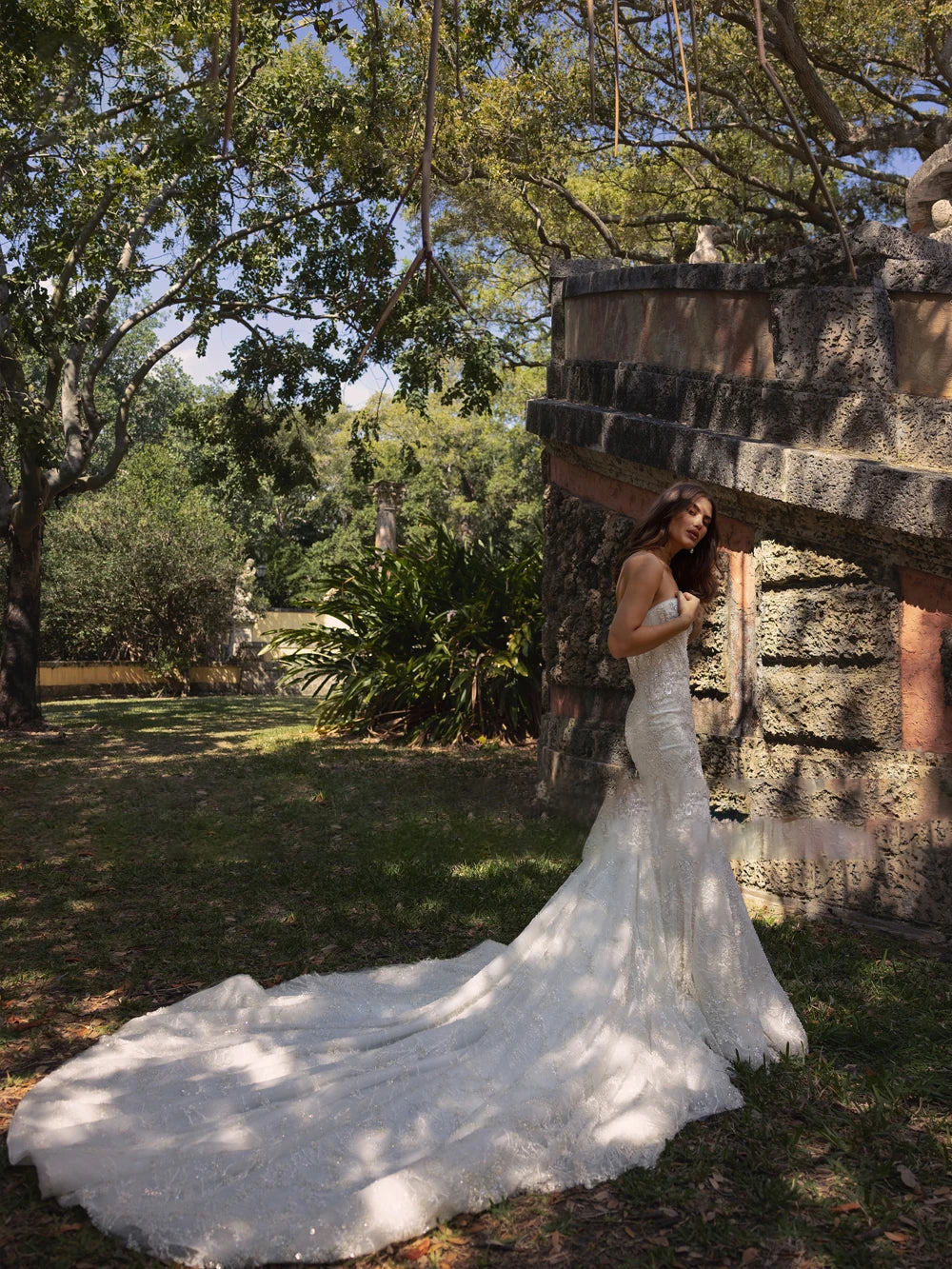 Romantic long sleeve lace wedding dress with subtle shimmer, emphasizing a structured corset bodice and statement feather wrist cuffs, styled leaning against a stone wall.